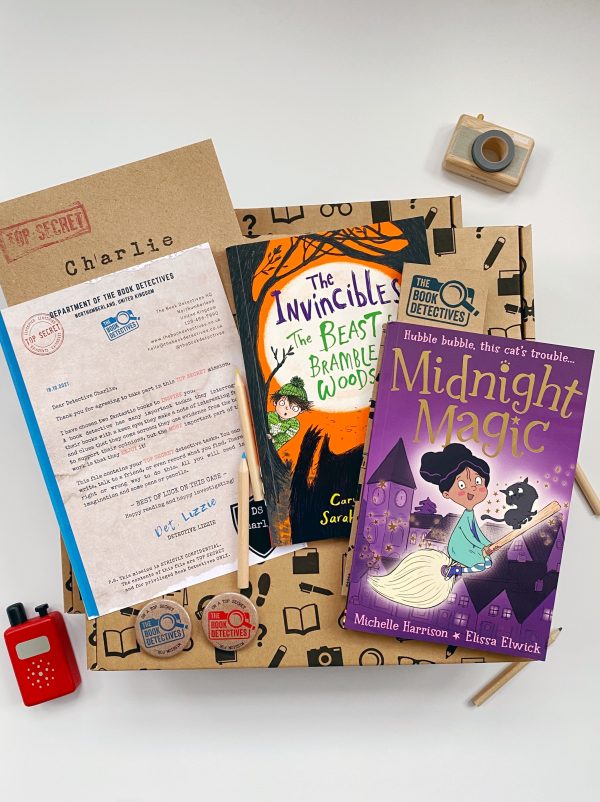 The image shows the contents of the book box laid out on a white background. There is a book with a purple cover and an illustration of a child riding a broomstick. The second book has an orange and black cover and shows an illustration of two children looking shocked at the shadow of a large cat. There is a letter written on a kraft background and a kraft envelope. There is a red walkie talkie, a wooden camera and some pencils for decoration.