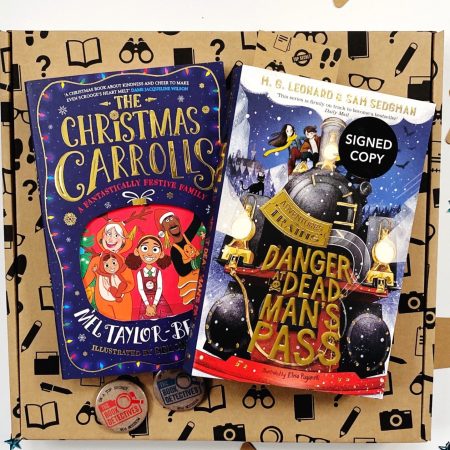 The image shows two books on top of a brown cardboard box. The books are The Christmas Carrolls and Danger At Dead Man's Pass. The books and box are on a white background and there are blue and brown stars for decoration placed around the box.