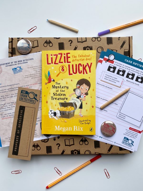 The image shows the contents of the book box. There is a brown cardboard box photographed, it has the book Lizzie and Lucky on top of it. There are activity sheets next to the box and some pencils, paper clips, badges, pens and a bookmark.